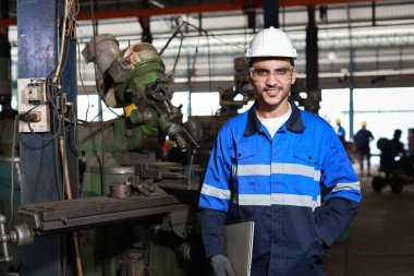 Koruyucu giysili mühendis ayakta ve bilgisayar kullanıyor ve kamerayı kullanıyor. Ağır sanayi imalat fabrikasında metal torna işi yapıyor.