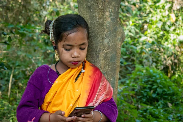 A tribal little girl is listening to music alone with Bluetooth headphones on her android phone in the forest. She is wearing traditional aboriginal attire.