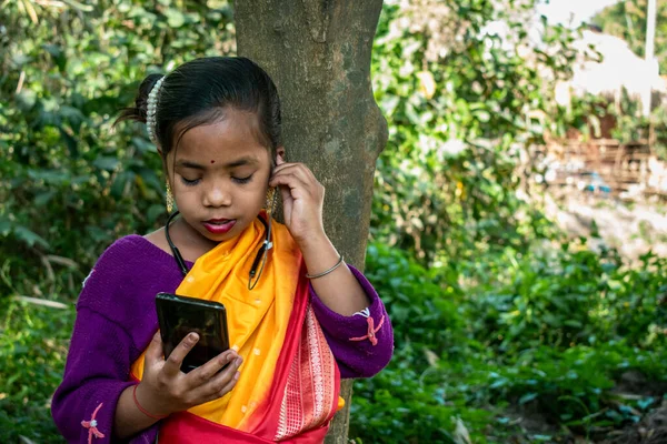 A tribal little girl is listening to music alone with Bluetooth headphones on her android phone in the forest. She is wearing traditional aboriginal attire.
