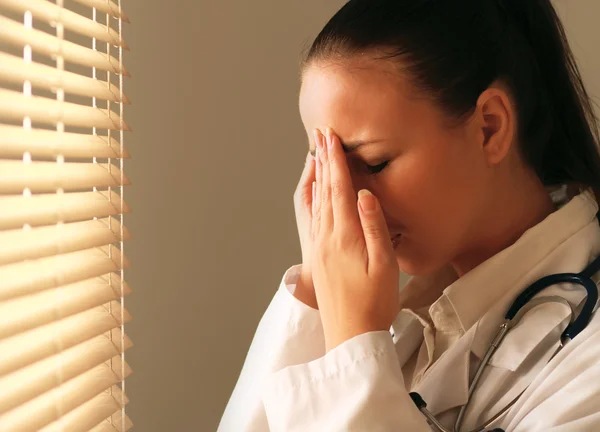 Médico femenino con dolor de cabeza de pie cerca de la ventana —  Fotos de Stock