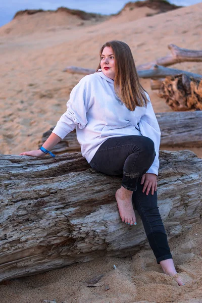 Blonde girl in black pants and white sweatshirt on the beach leaning on a stranded log.