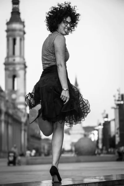 Woman winking with one eye and one leg raised in a city, black and white photograph. In Pilar square Zaragoza, Spain