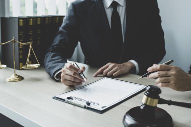 Avukatın eli kalem tutuyor ve ofisteki adama hukuki danışmanlık hizmeti veriyor. Adalet terazisi ve tokmak çekiciyle..