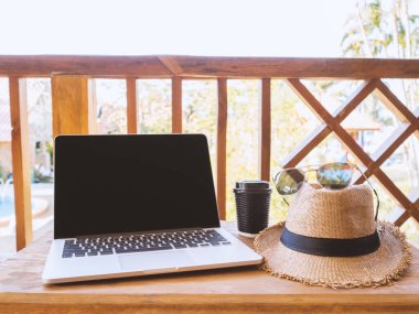 A laptop, coffee mug, hat, and eyeglasses sit on a brown wooden table.