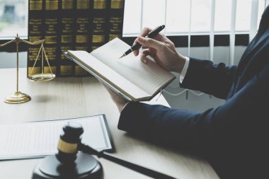 Avukatın eli kalem tutuyor ve ofiste hukuki danışmanlık hizmeti veriyor. Adalet terazisi ve tokmak çekiciyle..