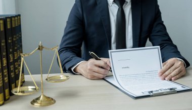 Avukatın eli kalem tutuyor ve ofiste hukuki danışmanlık hizmeti veriyor. Adalet terazisi ve tokmak çekiciyle..