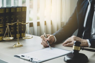 Avukatın eli kalem tutuyor ve ofiste hukuki danışmanlık hizmeti veriyor. Adalet terazisi ve tokmak çekiciyle..