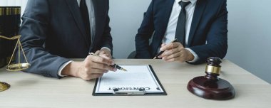 Avukatın eli kalem tutuyor ve ofisteki adama hukuki danışmanlık hizmeti veriyor. Adalet terazisi ve tokmak çekiciyle..