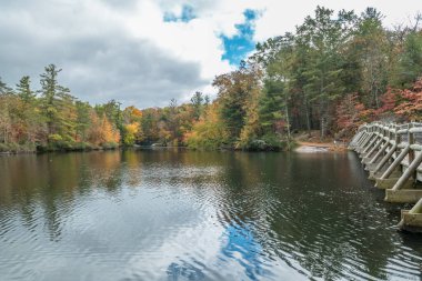 Ağaçların canlı renkleri gölde yansıyor. Sonbaharda bulutlu bir günde gölün etrafında yürüyüş izleri var.