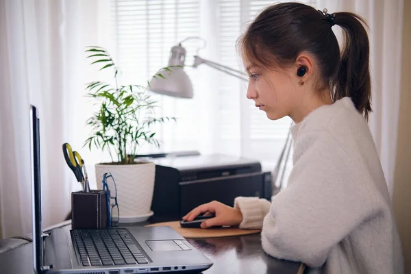 Joven hermosa chica trabaja en un ordenador portátil, sosteniendo una mano en un ratón de ordenador en casa en una mesa de trabajo en una habitación luminosa. — Foto de Stock