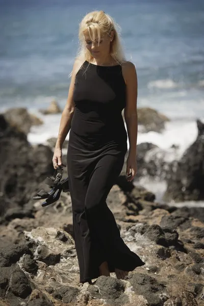 Vacker blond kvinna vandra längs steniga havsstrand i svart klänning medan du håller finskor — Stockfoto