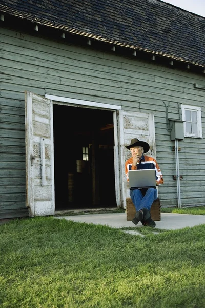 Homem de chapéu de cowboy trabalhando no computador fora do celeiro — Fotografia de Stock