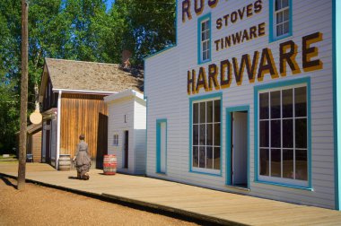 Woman In Period Dress Walking Past Historic Hardware Store At Fort Edmonton Park clipart