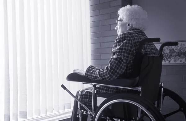 Senior vrouw zitten in rolstoel kijken uit venster met blinds — Stockfoto