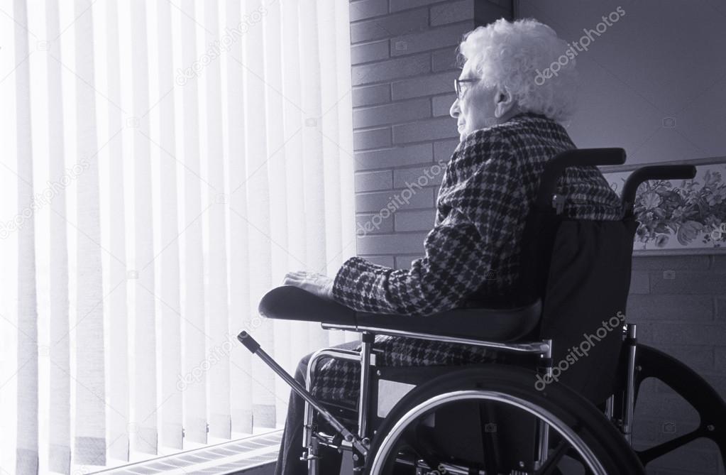 Senior Woman Sitting In Wheelchair Looking Out Of Window With Blinds