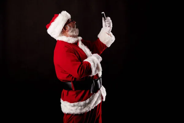 Noel Baba siyah arka planda cep telefonuyla selfie çekiyor.