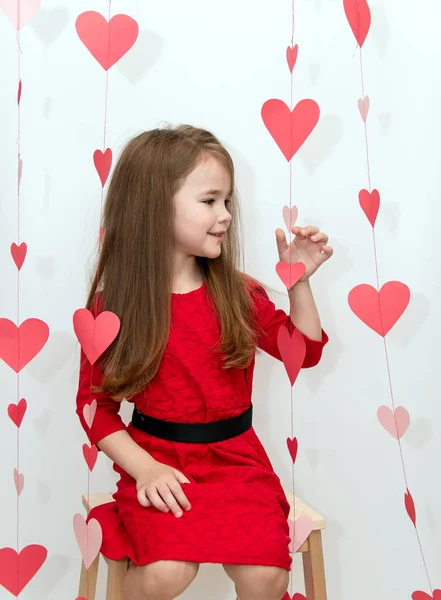 Девушка с длинными волосами в красном платье сидит на стуле на белом фоне с сердечками. День Святого Валентина. — стоковое фото