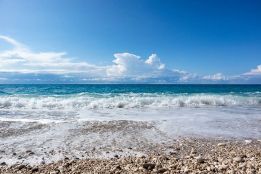 Güneşli fırtınalı dalgalar ve Yunanistan 'ın Lefkada adasının sahillerinde mavi suyu ve destansı bulutlu manzaralı beyaz çakıl taşlı plajda beyaz köpük. Yaz doğası İyon Denizi 'ne seyahat