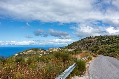 Yunanistan 'ın Lefkada adasındaki mavi deniz kıyısı yakınlarındaki yeşil tepelerde manzaralı bulutlu geleneksel Yunan köyü Kalamitsi' ye giden pitoresk yol. Yazın Avrupa seyahati
