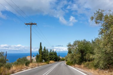 Yeşil zeytin ağaçları ve mavi gökyüzü manzaralı asfalt bir yol. Lefkada adasında deniz kıyısını dolaş, Yunanistan. Arabayla sahne gezisi