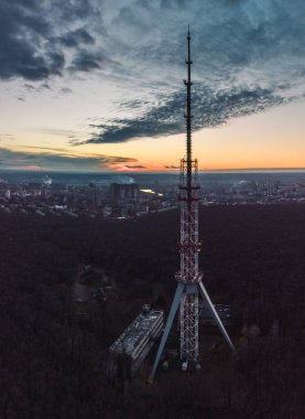 Karanlık bir ormanda, karanlık bulutlu gökyüzü olan telekomünikasyon kulesi anten silueti ile ormanın hava günbatımı dikey görüntüsü. Kharkiv, Ukrayna