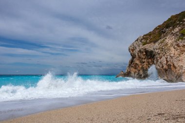 Beyaz köpüklü gök mavisi dalgalar kayalık kıyıları kırıyor. Lefkada adasında bulutlu gökyüzü olan Mylos kumlu plajı. Yaz doğası İyon Denizi 'ne seyahat