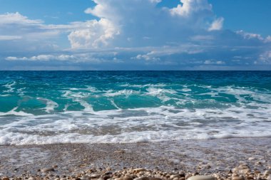 Yunanistan 'ın Lefkada adasının kıyısında mavi fırtınalı dalgalar ve destansı bulut manzaralı çakıl taşlı plajda manzaralı beyaz köpük. Yaz doğası İyon Denizi 'ne seyahat