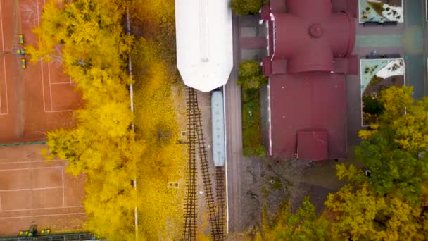 市内中心部の公園のテニスコートの近くにある子供南駅の上を飛んでください 鮮やかな黄色の秋の森の中で電車とプラットフォーム カラフルなハリコフの空中ツリートップビュー 旅行ウクライナ — ストック動画