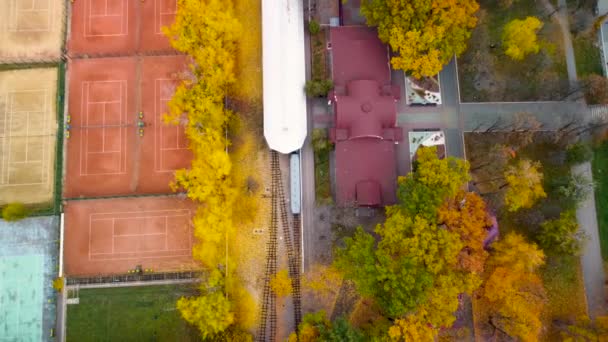 シティパークのテニスコート近くの子供南駅の上を後方に飛ぶ 鮮やかな黄色の秋の森の中でトラックラインと電車 カラフルなハリコフの空中ツリートップビュー 旅行ウクライナ — ストック動画