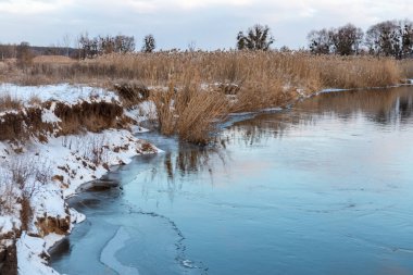 Soğuk nehirde kış manzarası bulutlu gökyüzü ayna suyunda yansıma ve kıyıdaki kuru sazlıklar. Nehir buz ve karla kaplı.