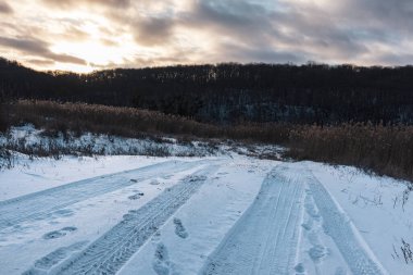 Kırsal bölgede lastik izleri olan karlı, beyaz kırsal bir yol. Akşam kış manzarası. Güneş, ağır bulutlar gökyüzü, sazlıklar ve karanlık ağaçlar orman tepesi.