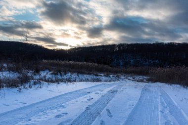 Kırsal bölgedeki karlı, beyaz kırsal yol. Akşam kış manzarası destansı ağır bulutlarda güneş, gökyüzü, sazlıklar ve orman tepeleri