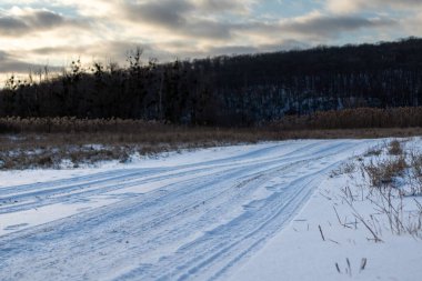 Kırsal kesimde, karlı, beyaz bir yol. Destansı ağır bulutlar gökyüzü, sazlıklar ve karanlık ağaçlar orman tepeleriyle akşam manzarası
