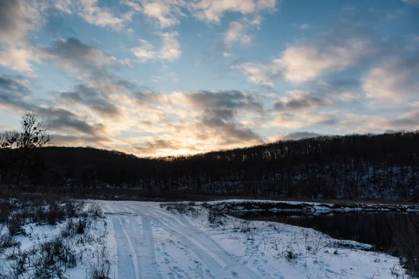 Kırsal bölgede lastik izleri olan karlı, beyaz kırsal bir yol. Akşam kış manzarası günbatımı güneşi destansı bulutlu gökyüzünde ve nehir kıyısında karanlık ağaçlar ormanında