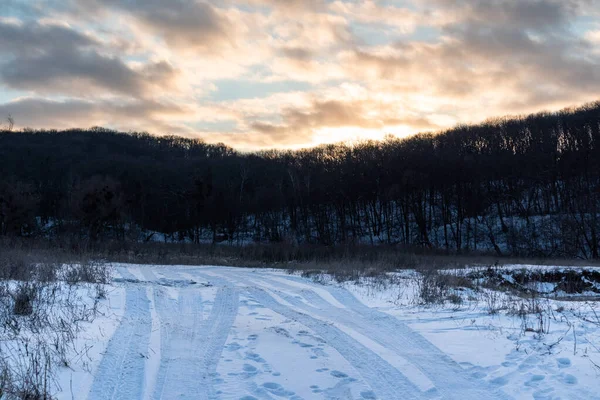 Kırsal bölgede lastik izleri olan karlı, beyaz kırsal bir yol. Akşam kış manzarası günbatımı güneşi destansı bulutlu gökyüzünde ve karanlık ağaçlar orman tepesinde.