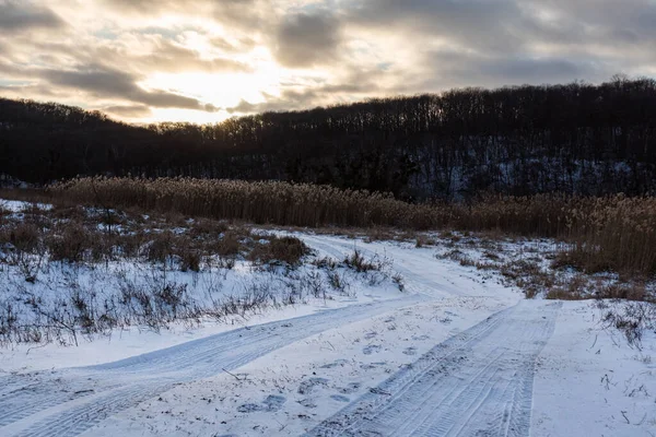 Kırsal bölgede kar üzerinde lastik izleri olan karlı, beyaz kırsal bir yol. Destansı ağır bulutlar, sazlıklar ve karanlık orman tepelerinde güneşli akşam manzarası
