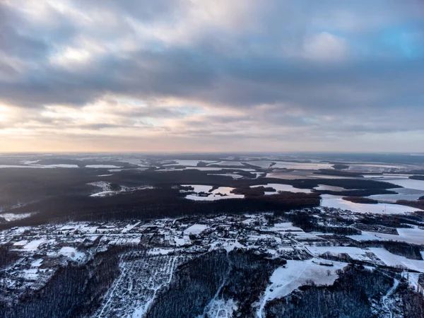 Köye kış destansı hava manzarası, karla kaplı orman ve tarlalar. Ukrayna 'nın Zmiyevsky bölgesi insansız hava aracından. Güneş, ağır gün batımı bulutlarında parlar.