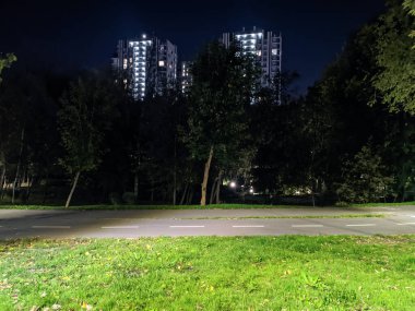 Modern konut binaları gece aydınlandı. Yeşil yaz şehir parkındaki bisiklet yolunun manzarası. Koyu ağaçlı siluetler.