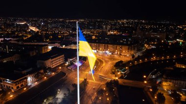 Ukrayna 'nın aydınlanmış bayrağı karanlık gece şehir manzarasında dalgalanıyor. Ukrayna 'nın başkenti Kharkiv' deki Skver Strilka yakınlarındaki Pavlivska Meydanı 'ndaki şehir trafiği hava görüntüsü