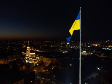 Illuminated yellow and blue flag of Ukraine waving on flagpole on dark evening sky. City lights aerial view with Holy Annunciation Cathedral in Kharkiv, Ukraine