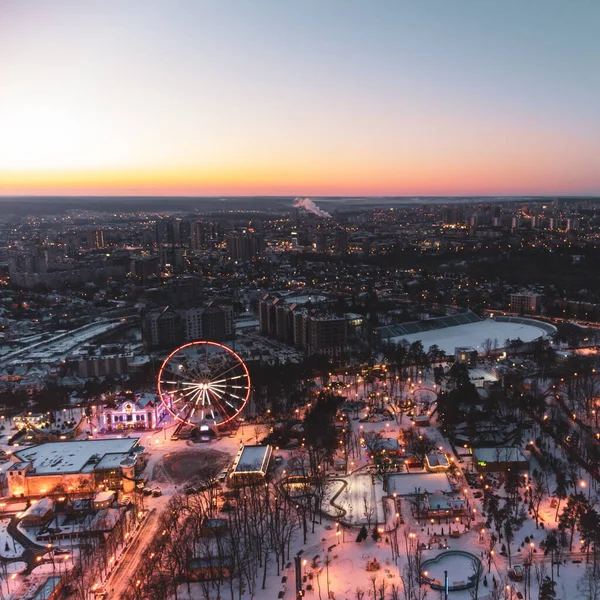 Canlı akşam şehri hava manzarası, dönme dolap ve kışın beyaz karla kaplı eğlenceler. Günbatımı renkleriyle Kharkiv şehir merkezi, eğlence Gorky Central Park