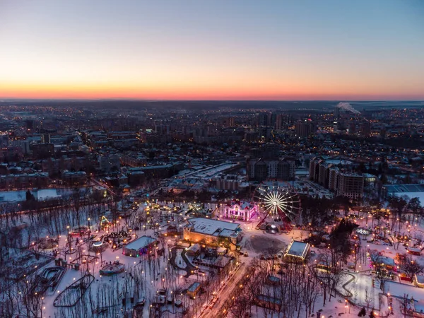 Canlı akşam şehir manzarası, dönme dolap ve kışın beyaz karla kaplı eğlenceler. Günbatımı renkleriyle Kharkiv şehir merkezi, eğlence Gorky Central Park