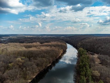 Aerial view on river curve in bare trees forest. Epic blue cloudscape reflection in water. Sunny nature landscape from drone