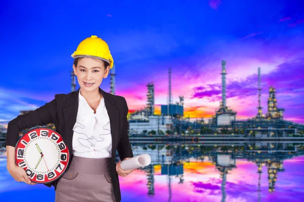 Μηχανικός γυναίκα σε κίτρινο κράνος, κρατώντας ένα μεγάλο ρολόι και το σχέδιο του έργου σε φόντο ενός διυλιστηρίου πετρελαίου — Φωτογραφία Αρχείου