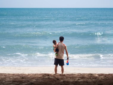 Baba ve çocuğu deniz sahilinde kuma bakıp kumla ve plastik oyuncakla oynamak için el sallıyorlar.