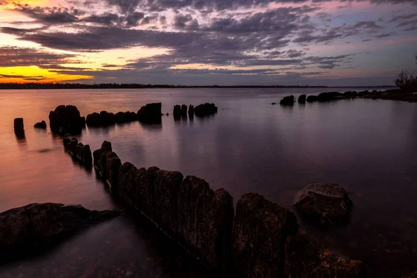 Beautiful sunset in the sky with clouds over the river. Lines of old wooden supports from the pier in the water in the foreground. Landscape at sunset