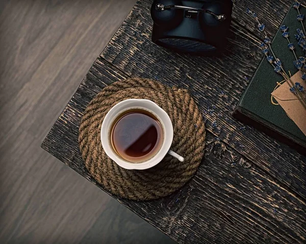 Bir fincan kahve, bir çalar saat, kurutulmuş lavanta filizi ve eski ahşap dokunun arka planında bir kitap..