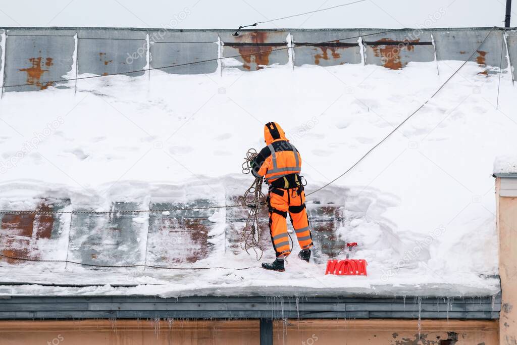 A working man in bright overalls with a safety belt with a shovel clears snow from the roof of an old building.