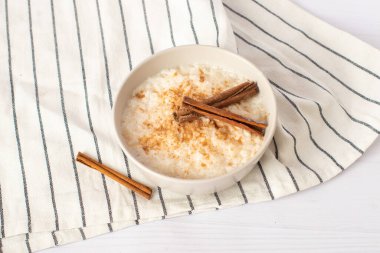 İskandinav usulü pirinç lapası. Beyaz arka plan. Sütlaç mavi bir seramik kasede, tahta bir masanın üzerinde, yukarıdan görülüyor. Zencefilli kurabiye ve tarçın çubuklarıyla