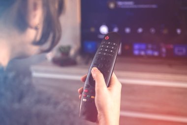 Uzaktan kumandayla televizyon seyreden bir kadın modern oturma odasındaki koltukta oturuyor.